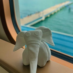 May include: A small, white elephant figurine with large ears and a trunk. The elephant is sitting on a brown surface, with a view of the ocean and a pier in the background. The figurine is made of a textured material.