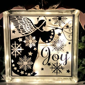 May include: A lit glass block featuring a black and white silhouette of an angel blowing a trumpet. The angel is surrounded by snowflakes and stars. The word "Joy" is written in a decorative font below the angel.