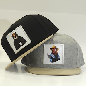 May include: Two baseball caps. One is black with a bear graphic holding a cup. The other is gray with a rooster graphic wearing a cowboy hat and a blue shirt. Both caps have a tan brim.