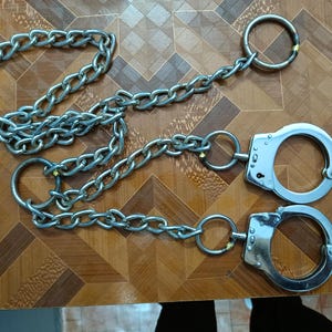 Puede incluir: Un par de esposas plateadas con cadenas de conexión y grandes anillos metálicos. Las esposas están sobre una superficie de madera marrón con un patrón geométrico. Las cadenas son de metal.