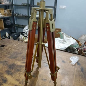 Könnte beinhalten: Ein Holzstativ im Vintage-Stil mit Messingbeschlägen. Das Stativ hat dunkelbraune Holzbeine und eine zentrale Messingsäule. Die Beine sind mit Messingstützen verbunden und haben Messingfüße. Die Oberseite hat einen Messinganschluss zur Montage von Geräten.