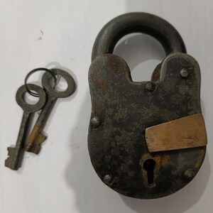 Puede incluir: Un candado antiguo y desgastado, con un acabado oscuro y envejecido. Dos llaves a juego están junto al candado. El candado tiene un ojo de cerradura y una placa metálica. El metal del candado tiene un aspecto rústico y desgastado.