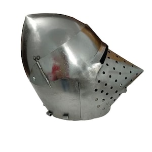 May include: A polished, silver-colored medieval helmet with a pointed visor and numerous ventilation holes. The helmet features a hinged visor and a curved, protective design, likely for historical reenactment or display.