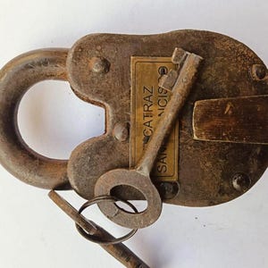 May include: A weathered, rusty padlock with two keys. The lock features a large, rounded shackle and a rectangular body. A small label on the lock reads "Alcatraz San Francisco". The keys are attached to a ring.