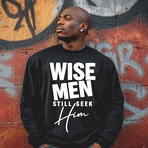 May include: Black sweatshirt with the text "WISE MEN STILL SEEK Him" in white. The sweatshirt is worn by a person with a shaved head, standing in front of a graffiti-covered wall with red, orange, and gray paint.