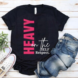 May include: Black t-shirt with the words "HEAVY" in pink vertically on the left side and "on the SELF Respect" in white. The shirt is paired with blue jeans and white sneakers, creating a casual, everyday look.