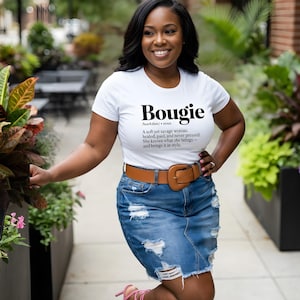 May include: White t-shirt with the word "Bougie" printed in black, along with a definition. The shirt is paired with a denim skirt and a brown belt with a large buckle. The woman is smiling.
