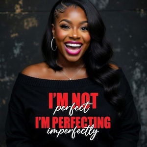 Pu&ograve; includere: Una donna indossa un maglione nero con spalle scoperte con la scritta "I'M NOT perfect I'M PERFECTING imperfectly" in rosso e bianco. Ha i capelli scuri, orecchini a cerchio e una collana. Sorride con un rossetto rosa acceso.