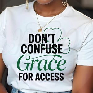 May include: White t-shirt with the message "DON'T CONFUSE Grace FOR ACCESS" in black and green lettering. A green shamrock design is next to the word "CONFUSE". The shirt is paired with blue jeans. A gold necklace and earrings are also visible.