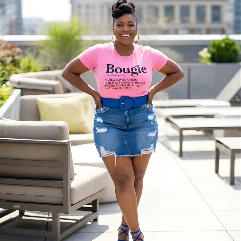以下が含まれることがあります： 「Bougie」の文字がプリントされたピンク色のTシャツを着た女性。ダメージ加工が施されたデニムスカートと青いベルトを着用。青いハイヒールも履いています。
