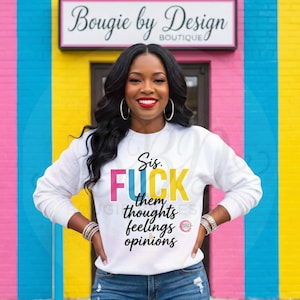 May include: A white sweatshirt with the text "Sis. FUCK them thoughts feelings opinions" in colorful block letters. The woman in the image is wearing the sweatshirt and smiling. The background is a colorful wall with the sign "Bougie by Design Boutique".