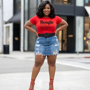 以下が含まれることがあります： 「Bougie」の文字が黒でプリントされた赤いTシャツを着た女性。デニムスカートと黒いベルトを合わせています。赤いストラップ付きのハイヒールも履いています。カジュアルでありながらファッショナブルなルックにスタイリングされています。