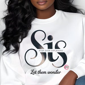 Könnte beinhalten: Weißes Sweatshirt mit dem Wort "Sis" in schwarzer und goldener Schrift. Der Text "Stop Over Explaining" steht über "Sis" und "Let them wonder" darunter. Ein kleines rosa Logo befindet sich in der unteren rechten Ecke.