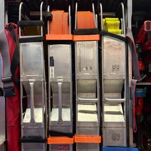 Könnte beinhalten: Ein Staufach mit vier vertikalen Metallgestellen für aufgerollte Feuerwehrschläuche. Die Gestelle verfügen über orangefarbene und gelbe Stoffgurte, silberne Metallgriffe sowie Beschriftungen wie DIN-14827-1 und Ziegler. Rote Ausrüstungstaschen liegen oben.