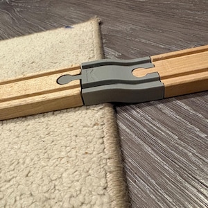 Könnte beinhalten: Holzspielzeug-Eisenbahnschiene mit einem grauen Kunststoffverbinder. Die Schiene ist hellbraun und verbindet sich mit einem anderen Teil. Die Schiene liegt auf einer Holzoberfläche und einem beigen Teppich.