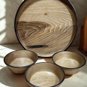 Op de afbeelding: Een houten serveerset met een grote ronde dienblad en drie kleine kommen. De dienblad en kommen hebben een lichte houtnerf afwerking met donkerbruine randen. De set is ontworpen voor het serveren van voedsel of het tentoonstellen van decoratieve items.