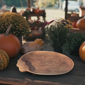 Può includere: Un vassoio ovale in legno chiaro con un dettaglio a forma di foglia. Sullo sfondo zucche, zucche ornamentali e fiori suggeriscono un tema autunnale. Il vassoio è probabilmente utilizzato per servire cibo o come elemento decorativo.