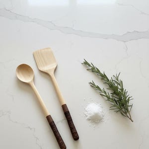May include: Wooden spoon and spatula with dark brown handles. A sprig of rosemary and a small pile of salt are next to the utensils. The kitchen tools are made of light-coloured wood.