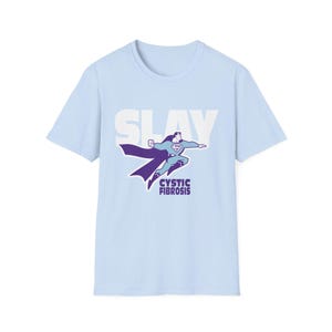 Puede incluir: Camiseta azul claro con un gráfico de un superhéroe volando, con la palabra "SLAY" arriba y "CYSTIC FIBROSIS" abajo. El superhéroe lleva un traje azul y morado con una capa.