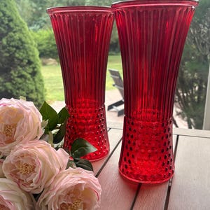 May include: Two tall, vibrant red glass vases featuring a vertical ribbed pattern on the upper section and a textured hobnail design on the lower base. A cluster of soft pink artificial peony flowers with green leaves sits beside the vases.