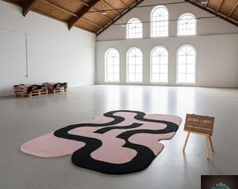 May include: A large, abstract rug with a pink base and a black, flowing design. The rug is displayed in a spacious, white-walled room with arched windows and a high, wooden ceiling. A small sign reads "Jaysha Carpet".