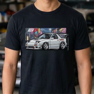 May include: A black t-shirt featuring a graphic illustration of a white sports car with pop-up headlights. The design includes Japanese text, the words Initial D, Red Suns, JDM Archives, and Tuning Techniques set against a city street background.