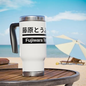 May include: A white travel mug with a black handle and a silver lid. The mug features Japanese text and the English text "Fujiwara To" on a black stripe. The mug is on a wooden table with a beach and ocean background.
