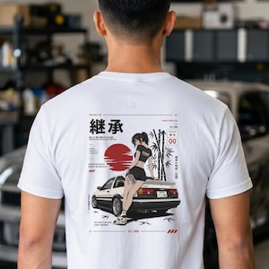 May include: A white t-shirt featuring a graphic on the back of a person leaning against a white and black sports car. The design includes a red sun, bamboo stalks, Japanese text, and English phrases like Inherited Tradition in Motion.