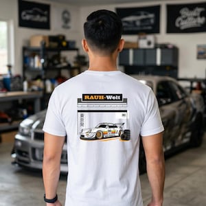 May include: White t-shirt with a graphic of a race car, the text "RAUH-Welt", and Japanese text. The car is white with yellow, orange, and black accents. A stack of tires is next to the car.