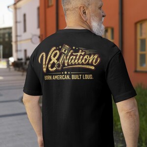 Könnte beinhalten: Ein schwarzes Kurzarm-T-Shirt mit einer Grafik auf der Rückseite, die den Text V8 Nation und eine Kolbenillustration zeigt. Unter dem Text ist der Schriftzug BORN AMERICAN. BUILT LOUD. in goldener Farbe mit einem Flaggenmuster gedruckt.