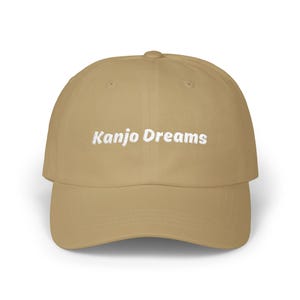 May include: A tan baseball cap featuring the white embroidered text "Kanjo Dreams" across the front. The cap has a curved peak and a rounded crown.