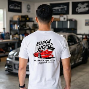 May include: White t-shirt with a graphic of a red sports car and the words "ROUGH WORLD" and "AIR/COOLED LEGENDS". The shirt also has the text "RWB 964 CLUB". The person is standing in a garage with cars.