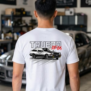 May include: A white short-sleeve t-shirt featuring a graphic on the back of a black and white Toyota AE86 Trueno car. The text "TRUENO" is printed in large black letters above the car, with "AE86" in red script to the right of the car graphic.