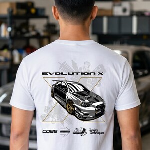 Peut inclure: Un t-shirt blanc avec un motif au dos représentant une voiture Mitsubishi Evolution X argentée aux jantes dorées. Le design comprend le texte "EVOLUTION X" au-dessus de la voiture, une grande forme en "X", une silhouette urbaine et des logos.