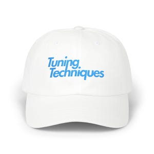 May include: White baseball cap with the text "Tuning Techniques" in blue script across the front. The cap has a curved brim and a classic six-panel design. A casual accessory for everyday wear.