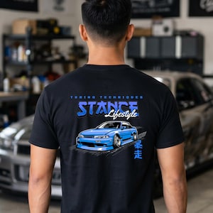 May include: Black t-shirt with a blue car graphic and the words "TUNING TECHNIQUES STANCE Lifestyle". The car is a light blue coupe with a lowered suspension. The Japanese characters on the right side of the car are also in blue.