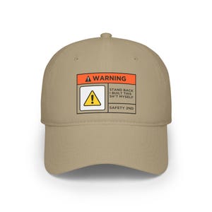 Op de afbeelding: Beige baseballpet met een waarschuwingslabel. Het label toont een gevaarsymbool en de tekst "WARNING", "STAND BACK I BUILT THIS SH*T MYSELF SAFETY 2ND".