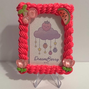 May include: A pink picture frame with a decorative, textured border. The frame is adorned with fruit-themed embellishments, including cherries, a watermelon slice, and flowers. The frame holds a card with a cloud and fruit design, and the text "DreamBerry NYC".