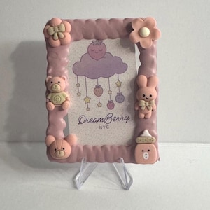 May include: Pink rectangular picture frame with a textured border, decorated with a teddy bear, bunny, flower, and baby bottle. The frame has a printed design with a cloud, strawberry, and the text "DreamBerry NYC".