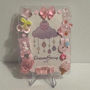 May include: A clear acrylic display featuring pink, translucent charms in various shapes, including a bow, stars, cherries, and bunnies. The center has a design with a cloud, strawberries, and the text "DreamBerry NYC."
