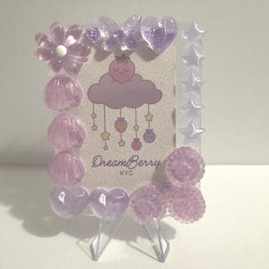 May include: A decorative picture frame with a light purple and white color scheme. The frame is adorned with various embellishments, including flowers, seashells, butterflies, hearts, and stars. The frame holds a card with a cloud and the text "DreamBerry NYC".