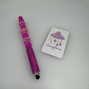 May include: A pink stylus pen with a sparkling, rhinestone-covered barrel and a series of pink and crystal-like beads. A small card with the text "DreamBerry NYC" and a cloud illustration is next to the pen.