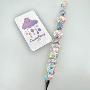 May include: A ballpoint pen decorated with iridescent pastel beads and small seashell charms, accompanied by a white rectangular card featuring a purple cloud, strawberry, and star graphic with the text DreamBerry NYC printed in black.