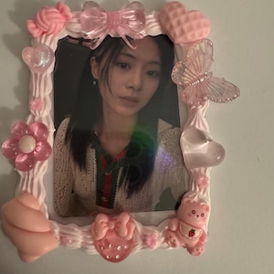 May include: A decorative photo frame with a picture of a person. The frame is white with pink embellishments, including a bow, hearts, a butterfly, a flower, and a bear. The frame has a textured, whipped cream-like design.
