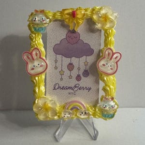 May include: Yellow resin picture frame with bunny and cupcake accents. The frame features a DreamBerry NYC print with a rainbow and cloud design. The frame is supported by a clear plastic stand.