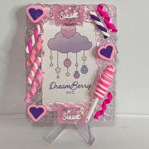 May include: A pink picture frame with a decorative border, featuring a central image with a cloud and hanging fruit design. Embellishments include pink hearts, candy swirls, and a lollipop. The frame has the text "Sweet" and "DreamBerry NYC".