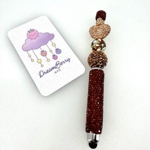 May include: A pen covered in dark red rhinestones with a decorative topper featuring a heart-shaped bead, a metallic sphere, and a smaller dark red bead. Beside it is a white rectangular card with the text DreamBerry NYC and a purple cloud graphic.