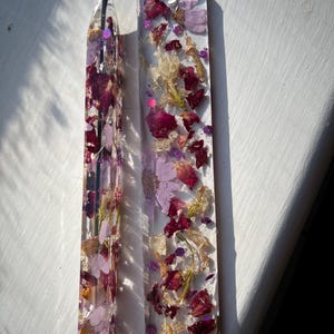 Könnte beinhalten: Zwei Lesezeichen und ein Stift aus Kunstharz, gefüllt mit getrockneten Blumen in Rot-, Lila- und Gelbtönen. Der Stift hat eine schwarze Tintenpatrone und eine rote Spitze. Die Lesezeichen sind rechteckig und haben oben ein Loch für eine Quaste.
