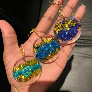 Può includere: Tre portachiavi rotondi in resina trasparente con elementi dorati. Ogni portachiavi presenta un design circolare riempito di glitter colorati. I glitter includono sfumature di blu, oro e verde acqua, creando un effetto scintillante. I portachiavi sono tenuti in una mano.