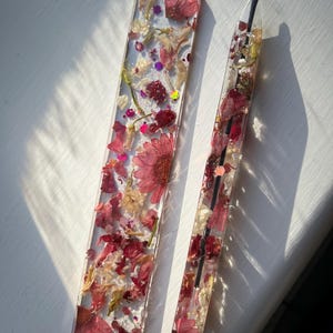 May include: A clear resin bookmark and pen set. The bookmark is rectangular, and the pen is a similar shape. Both are filled with dried red and cream-coloured flowers, glitter, and small purple accents. The pen has a black ink cartridge.
